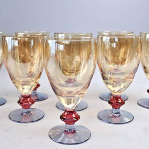 May include: A collection of eight vintage wine glasses. Each glass has a clear, amber-tinted bowl, a red stem, and a blue base. The glasses are displayed on a white surface, suitable for a formal setting.
