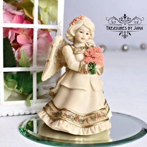 Puede incluir: Una figurita de porcelana de un ángel con un vestido blanco y flores rosas. El ángel tiene un halo y alas. La figurita está sobre una base de espejo.