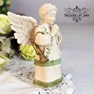 Puede incluir: Una figura de ángel color crema con las manos juntas y los ojos cerrados, vestida con una túnica blanca con ribetes verdes y sosteniendo una trompeta dorada. El ángel tiene alas grandes y detalladas y está sobre una superficie blanca. El texto "Treasures by Jana" es visible.