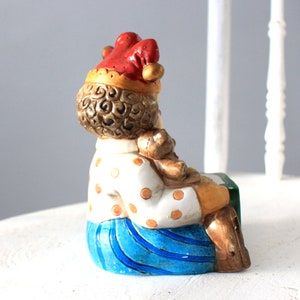 May include: A ceramic figurine of a clown holding a teddy bear. The clown is wearing a red and white hat, a white shirt with brown polka dots, and blue trousers. The figurine is sitting on a green and white base.