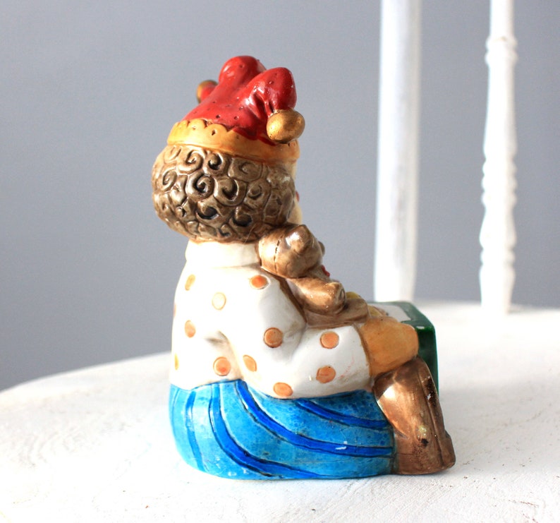 May include: A ceramic figurine of a clown holding a teddy bear. The clown is wearing a red and white hat, a white shirt with brown polka dots, and blue trousers. The figurine is sitting on a green and white base.