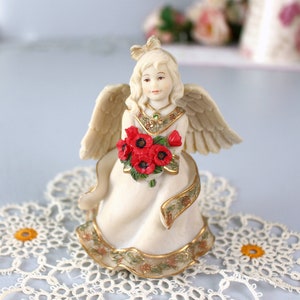 Puede incluir: Figura de ángel color crema con un ramo de amapolas rojas. El ángel tiene alas detalladas, un lazo y un vestido decorativo con adornos dorados. La figura está colocada sobre un tapete de encaje blanco.