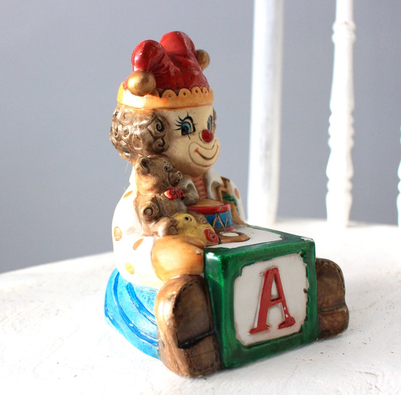 May include: A ceramic clown figurine wearing a red and white hat, holding a teddy bear and a toy drum. The clown is sitting on a green block with the letter 'A' on it.