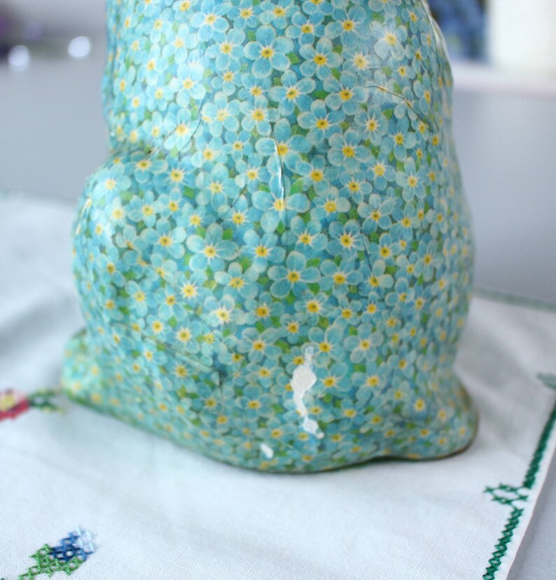 Pode incluir: Uma figurinha de cer&acirc;mica de um coelho coberta com um padr&atilde;o floral azul e amarelo. O coelho est&aacute; sentado em uma superf&iacute;cie branca.