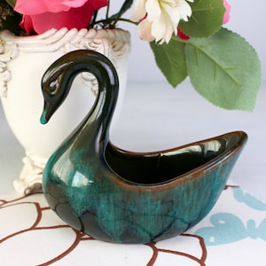May include: A ceramic swan-shaped planter with a glossy, teal and brown glaze. The swan has a long, curved neck and a small, open basin for holding plants or other decorative items. The planter is a vintage piece.