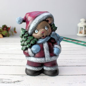 May include: A ceramic Christmas bear figurine wearing a red Santa suit with white trim, a blue scarf, and blue mittens. The bear is holding a small green Christmas tree.