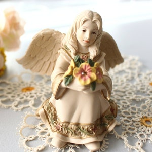 Puede incluir: Una figura de ángel color crema con alas extendidas, sosteniendo un ramo de flores amarillas y rosas. El vestido del ángel está adornado con detalles florales y dorados. La figura está arrodillada, con una expresión serena.