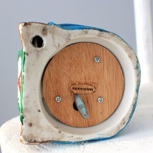 May include: A white ceramic musical figurine with a blue, green and red painted design. The figurine has a wooden back panel with a winding key and the word "INNOVATION" printed on it.