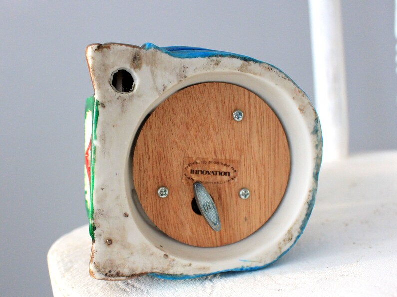 May include: A white ceramic musical figurine with a blue, green and red painted design. The figurine has a wooden back panel with a winding key and the word "INNOVATION" printed on it.