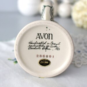 Op de afbeelding: Een witte keramische mok met een groene handgreep en een bloemenontwerp. De onderkant van de mok heeft de tekst "AVON Handcrafted in Brazil exclusively for Avon Products, Inc., 1983 286801."