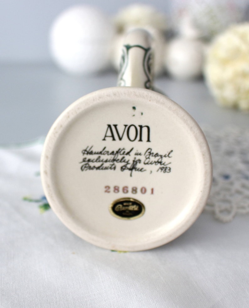 Op de afbeelding: Een witte keramische mok met een groene handgreep en een bloemenontwerp. De onderkant van de mok heeft de tekst "AVON Handcrafted in Brazil exclusively for Avon Products, Inc., 1983 286801."
