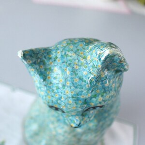 Pode incluir: Uma figurinha de cer&acirc;mica azul de um gato com um padr&atilde;o de flores de miosotis. O gato est&aacute; sentado com a cabe&ccedil;a inclinada para baixo.