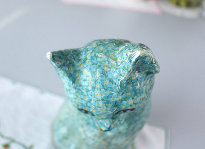 Pode incluir: Uma figurinha de cer&acirc;mica azul de um gato com um padr&atilde;o de flores de miosotis. O gato est&aacute; sentado com a cabe&ccedil;a inclinada para baixo.