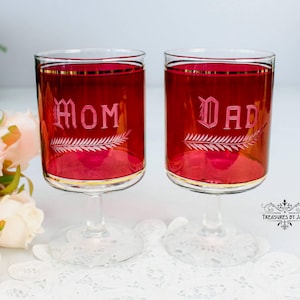 Peut inclure: Deux verres à vin vintage avec une teinte rouge, une bordure dorée et les mots "Mom" et "Dad" gravés dans une écriture élégante. Chaque verre présente un motif de fougère décoratif. Les verres sont remplis d'un liquide rouge.