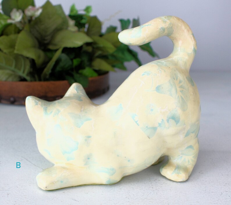 K&ouml;nnte beinhalten: Keramik-Katzenfigur in spielerischer Pose, bemalt in Cremegelb mit hellblauen floralen Akzenten. Die Katze ist gew&ouml;lbt, der Schwanz nach oben gebogen. Die Figur ist ein dekorativer Artikel.