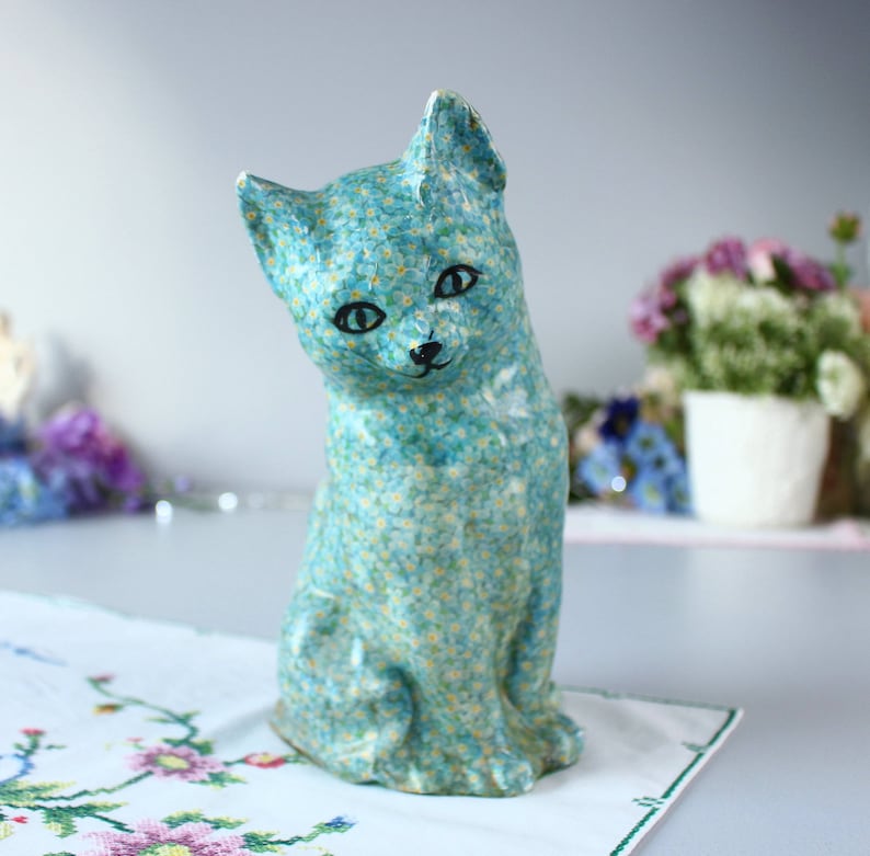 May include: A blue ceramic cat figurine with a forget-me-not flower pattern. The cat is sitting with its head tilted to the side and its eyes closed.