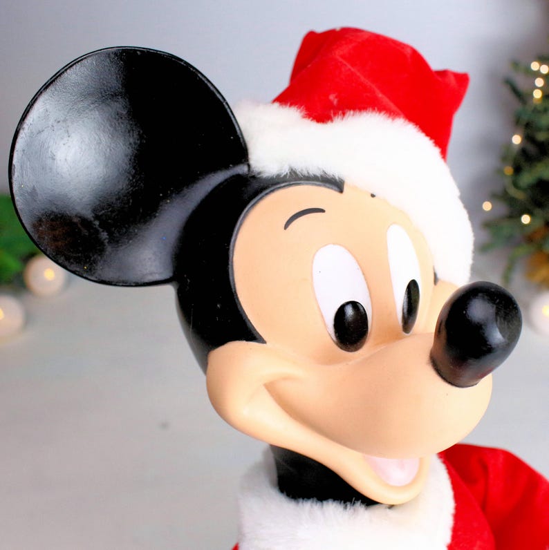 May include: A plush Mickey Mouse wearing a red Santa hat with white trim. The toy has black ears, black eyes, and a big smile.