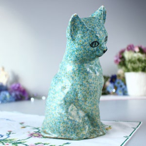 Pode incluir: Uma figura de gato de cer&acirc;mica azul com um padr&atilde;o de flores de miosotis. O gato est&aacute; sentado com a cabe&ccedil;a inclinada para o lado.