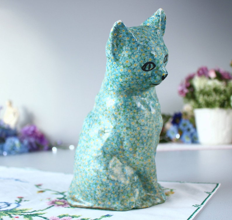 Pode incluir: Uma figura de gato de cer&acirc;mica azul com um padr&atilde;o de flores de miosotis. O gato est&aacute; sentado com a cabe&ccedil;a inclinada para o lado.