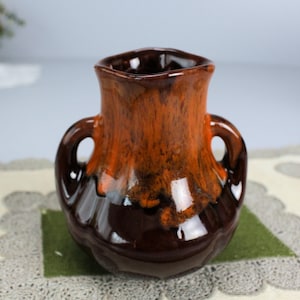 May include: A small, brown and orange ceramic vase with two handles. The vase has a mottled glaze and a rounded shape.