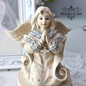 Puede incluir: Figura de ángel color crema con las manos juntas y un collar de cruz plateada. El ángel tiene cabello rubio claro, alas y viste una túnica con detalles azules y patrones de estrellas. El texto "Treasures by Jana" es visible.