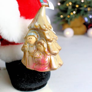 May include: A ceramic Christmas tree ornament featuring a depiction of the nativity scene. The ornament is beige with a brown glaze and depicts Mary, Joseph, and baby Jesus.