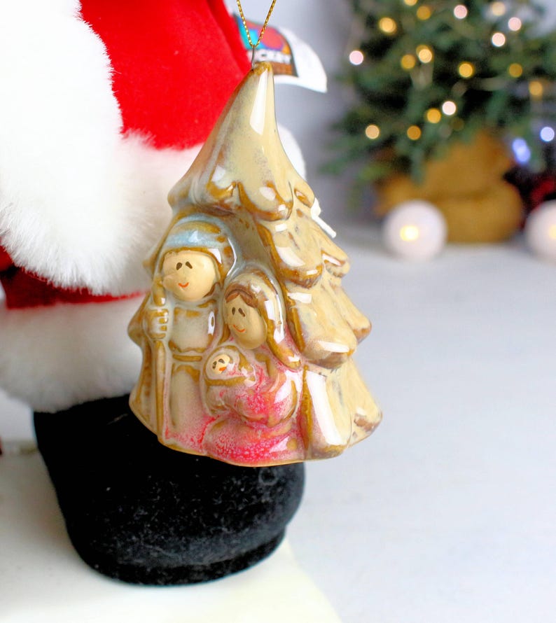 May include: A ceramic Christmas tree ornament featuring a depiction of the nativity scene. The ornament is beige with a brown glaze and depicts Mary, Joseph, and baby Jesus.