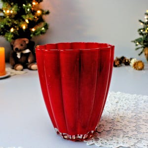 Könnte beinhalten: Eine rote Glasvase mit geriffeltem Design. Die Vase steht auf einer weißen Spitzentischdecke.
