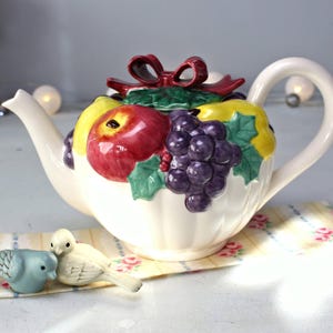 May include: A white ceramic teapot with a fruit design. The teapot is decorated with grapes, apples, pears, and a red bow. The teapot is sitting on a floral patterned fabric.