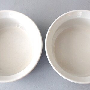 May include: Two white ceramic bowls with a simple, rounded design. The bowls are empty and have a smooth, glossy finish. The bowls are set against a light grey background. Ideal for serving soups, salads, or desserts.