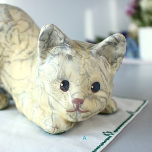 K&ouml;nnte beinhalten: Eine cremefarbene Keramik-Katzenfigur mit grauem Blumenmuster. Die Katze hat blaue Augen und eine rosa Nase und liegt auf einem wei&szlig;en Tuch mit gr&uuml;nem Stickrand. Der Buchstabe "A" ist sichtbar.