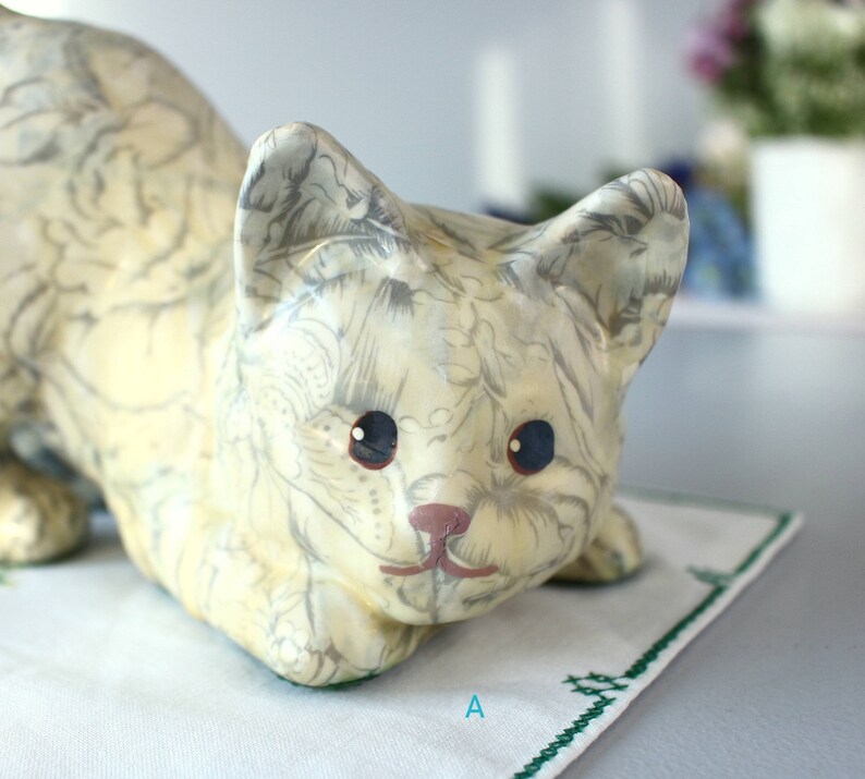 K&ouml;nnte beinhalten: Eine cremefarbene Keramik-Katzenfigur mit grauem Blumenmuster. Die Katze hat blaue Augen und eine rosa Nase und liegt auf einem wei&szlig;en Tuch mit gr&uuml;nem Stickrand. Der Buchstabe "A" ist sichtbar.