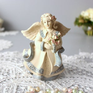 Puede incluir: Una figura de ángel color crema que sostiene un ramo de rosas rosas. El ángel tiene alas detalladas y viste una túnica azul claro y crema con adornos decorativos. Un diseño en forma de cinta fluye alrededor de la base de la figura.