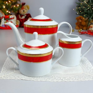 May include: A white porcelain tea set with red and gold accents. The set includes a teapot, sugar bowl, and creamer. The teapot has a curved spout and a lid with a small knob. The sugar bowl has a lid and two handles. The creamer has a handle and a pouring spout.