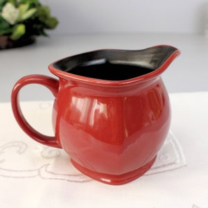 Peut inclure: Une crémière en céramique rouge brillant avec un intérieur noir. La crémière a un corps arrondi, une anse incurvée et un bec verseur. Le design est simple et élégant, adapté pour servir de la crème ou d'autres liquides.