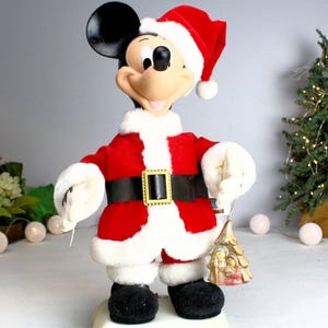 May include: A vintage animated Mickey Mouse Christmas figurine dressed in a red Santa suit with a white fur trim, holding a gold ornament with a nativity scene.