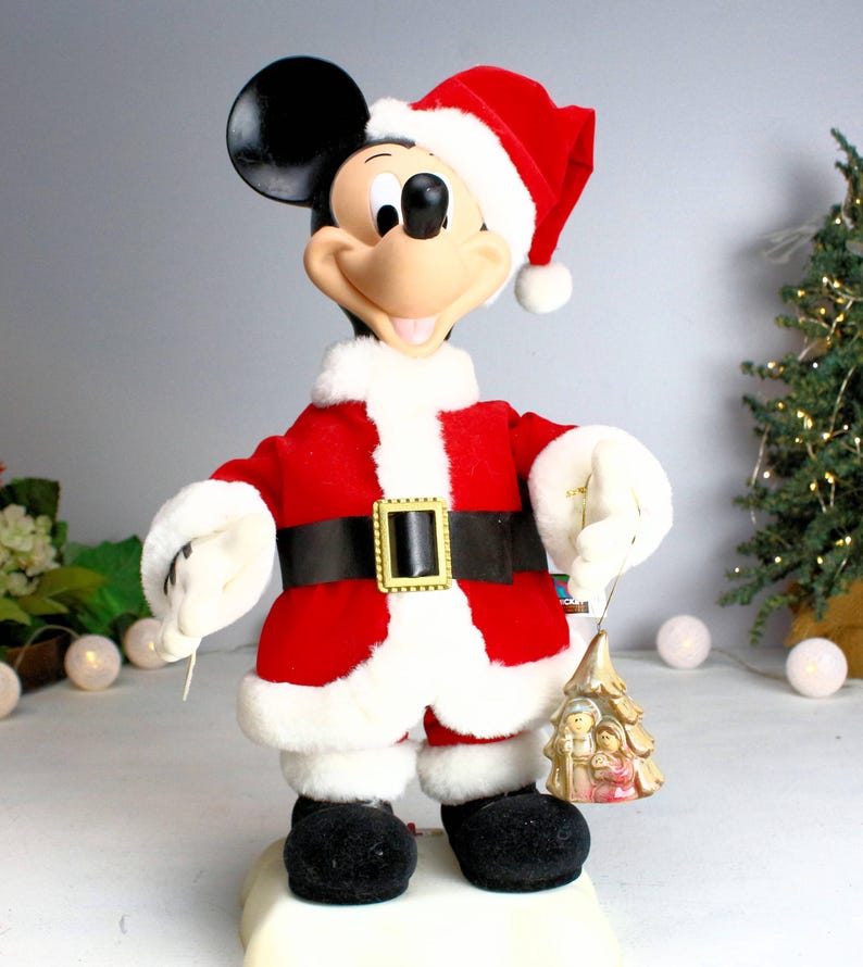 May include: A vintage animated Mickey Mouse Christmas figurine dressed in a red Santa suit with a white fur trim, holding a gold ornament with a nativity scene.
