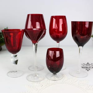 May include: Set of six red wine glasses, including a martini glass, a goblet, and four wine glasses. The glasses are all made of clear glass with a red tint.