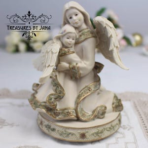 Puede incluir: Figurilla de ángel color crema que representa a un ángel adulto abrazando a un ángel niño. Los ángeles tienen alas emplumadas y visten túnicas fluidas con adornos dorados y verdes. La base de la figurilla está decorada con motivos florales. El texto "Treasures by Jana" es visible.