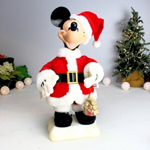 May include: A plush Mickey Mouse dressed as Santa Claus, wearing a red suit with white trim and a Santa hat. He is holding a small gold Christmas tree ornament.