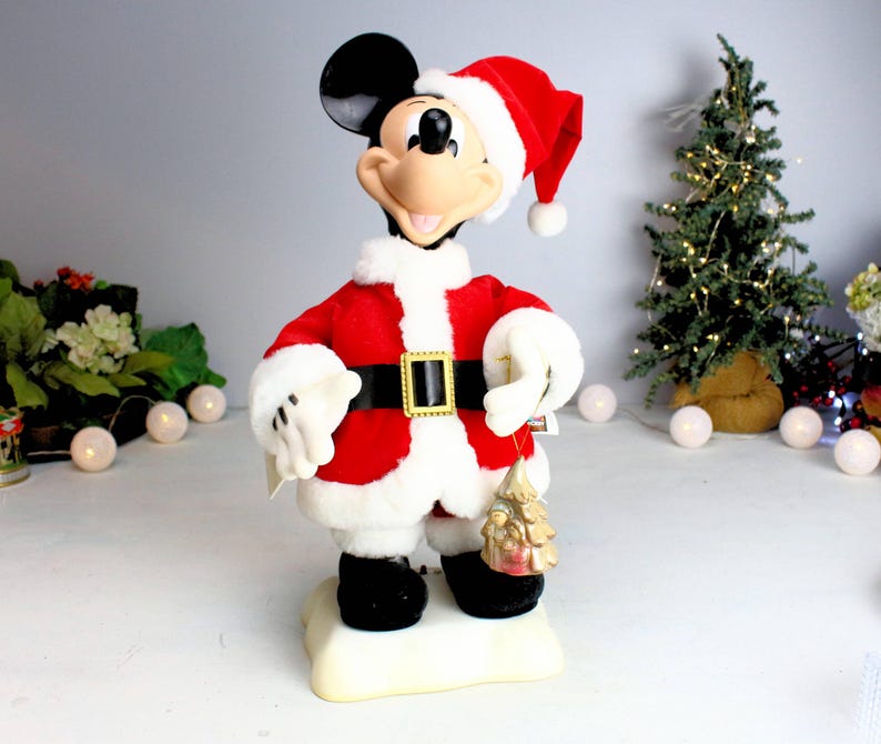 May include: A plush Mickey Mouse dressed as Santa Claus, wearing a red suit with white trim and a Santa hat. He is holding a small gold Christmas tree ornament.