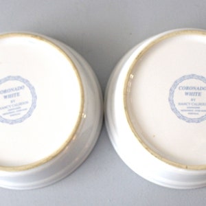 May include: Two white ceramic bowls with a slightly raised rim. The base of each bowl has a blue circular stamp with the text "CORONADO WHITE BY NANCY CALHOUN". These bowls are suitable for serving or display.