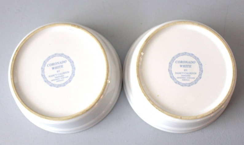 May include: Two white ceramic bowls with a slightly raised rim. The base of each bowl has a blue circular stamp with the text "CORONADO WHITE BY NANCY CALHOUN". These bowls are suitable for serving or display.