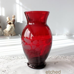 Peut inclure: Un vase en verre rouge rubis avec un motif floral gravé. Le vase a un dessus évasé et une base sombre. Le mot "etched" est imprimé en bas. Le vase est posé sur une surface blanche.