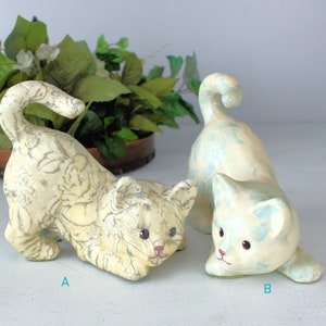 K&ouml;nnte beinhalten: Drei Keramik-Katzenfiguren. Eine Katze ist cremefarben mit floralem Muster, die anderen sind hellblau und cremefarben. Die Katzen befinden sich in spielerischen Posen, mit gebogenen Schw&auml;nzen und gesenkten K&ouml;pfen. Die Figuren haben blaue Augen und rote Nasen.