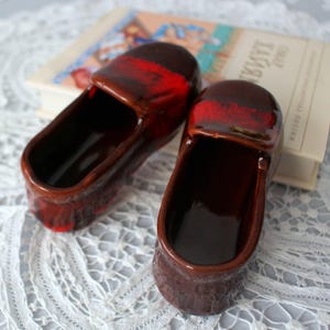 May include: A pair of miniature ceramic shoes in a deep brown hue with a glossy finish. Each shoe has a red band across the top. They are displayed on a white lace surface, with a book visible in the background.