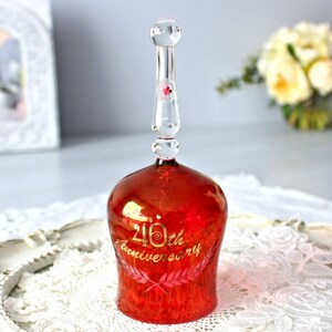 Peut inclure: Une cloche en verre rouge avec une poignée en verre transparent. La cloche est décorée d'une inscription dorée "40th Anniversary" et d'une couronne rose.