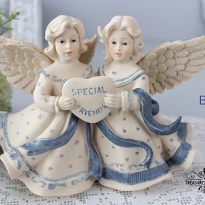 Puede incluir: Figura de ángel color crema con dos figuras sosteniendo un letrero en forma de corazón que dice "SPECIAL FRIENDS" en letras azules. Los ángeles visten vestidos fluidos con detalles azules y alas emplumadas. El objeto es decorativo.