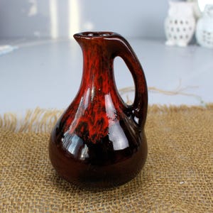 May include: A small, red and black ceramic jug with a rounded body and a handle. The jug has a glossy finish and a mottled glaze.