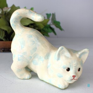 K&ouml;nnte beinhalten: Cremefarbene Katzenfigur mit hellblauem Blumenmuster. Die Katze befindet sich in einer spielerischen Pose, mit gebogenem Schwanz und gew&ouml;lbtem K&ouml;rper. Die Katze hat eine rote Nase und schwarze Augen.